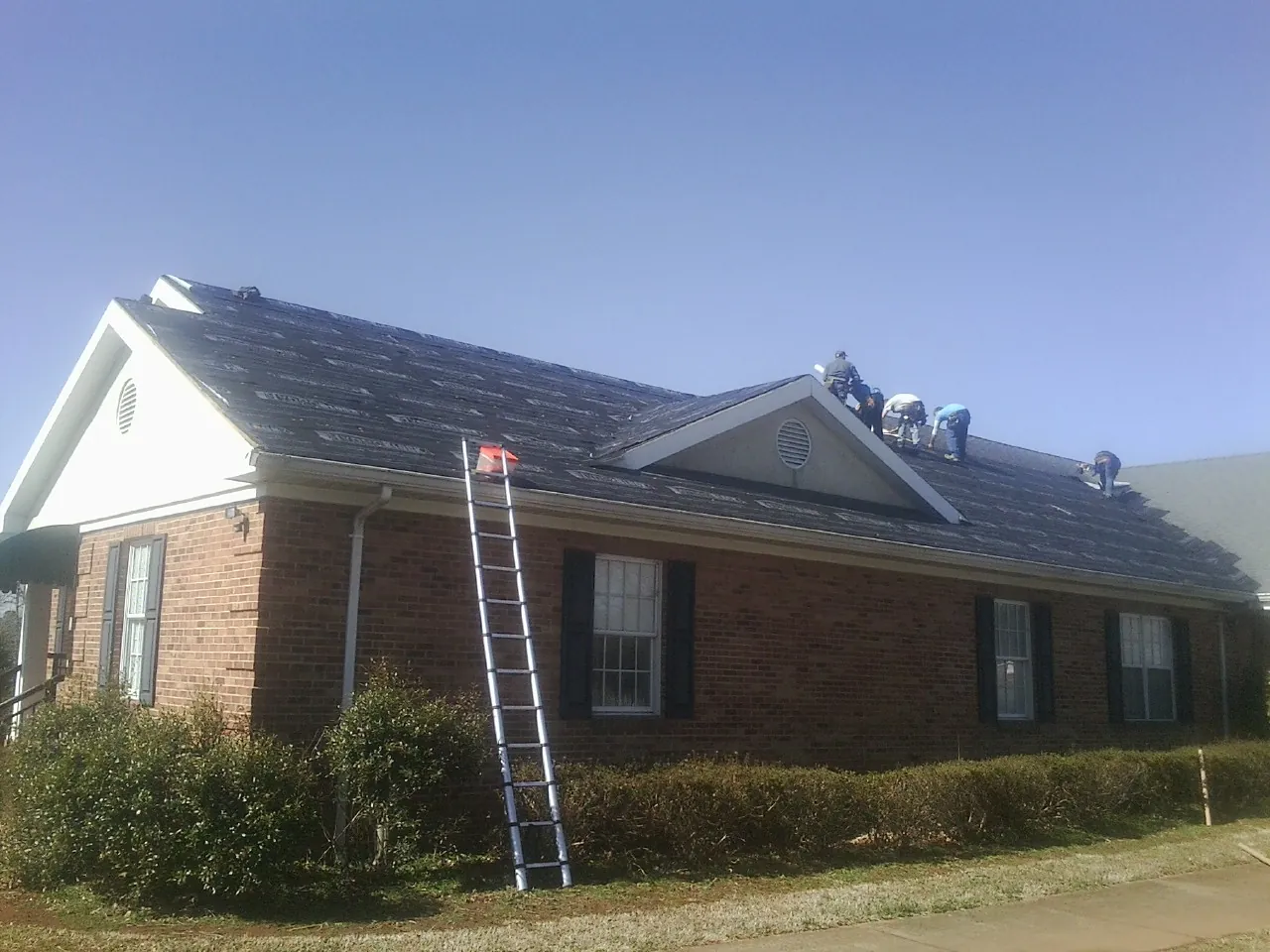 Crew tearing shingles off a brick ranch roof with a ladder set — Emergency Roof Repair work by our crew in May
