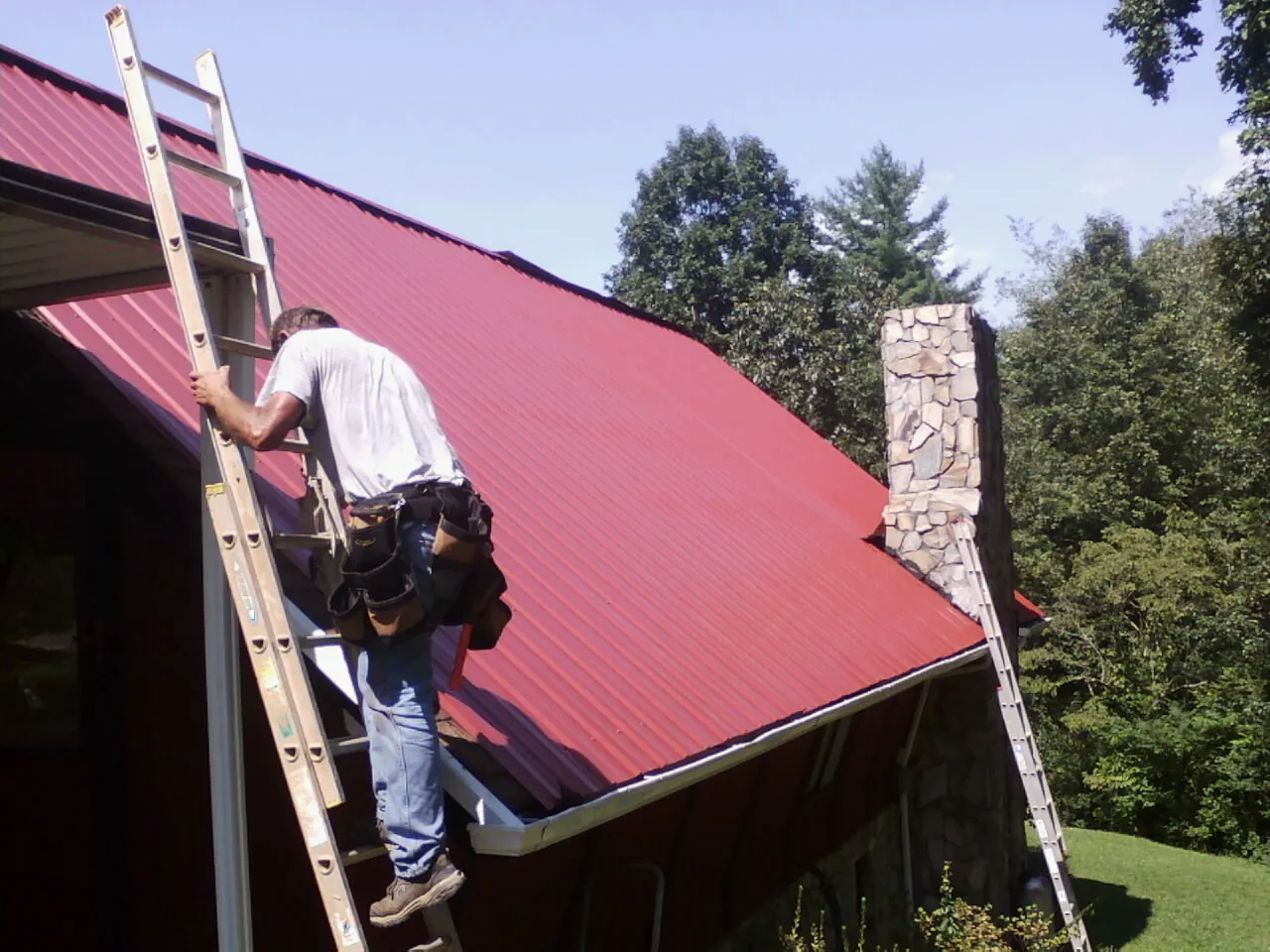 Roofer on a ladder beside a red metal roof and stone chimney — Soffit & Fascia Repair by Roofing Brothers in May